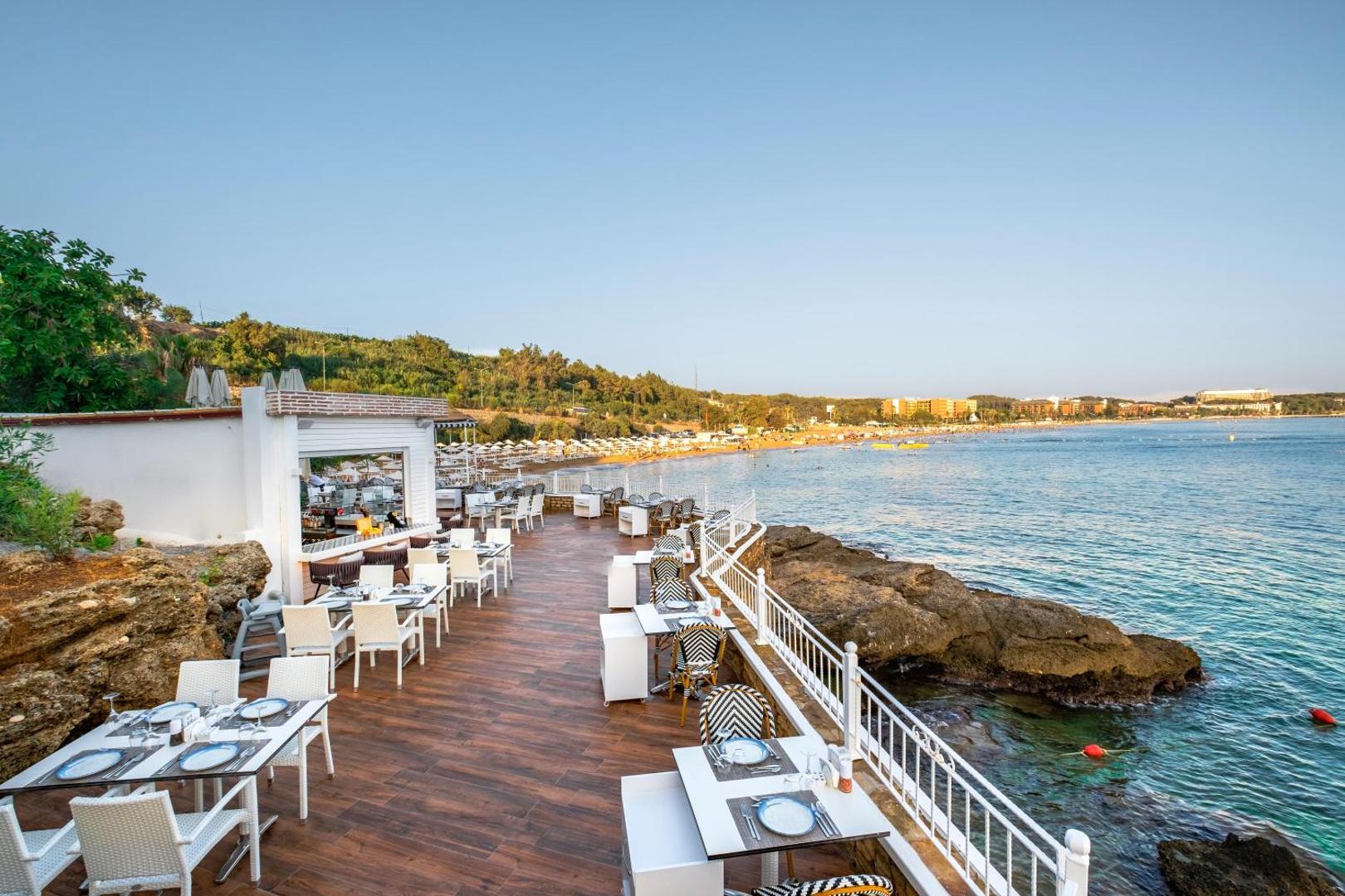 Utopia Beach Club āra terases restorāns uz klints piekrastē ar skatu uz jūru un kaimiņu pludmali saulrietā