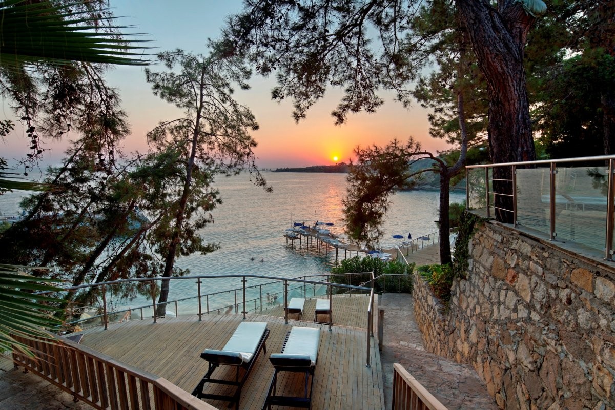 Utopia Beach Club koka terase ar sauļošanās krēsliem Alanjas piekrastē saulrieta laikā — skats uz jūru un koka piestātni caur priedēm