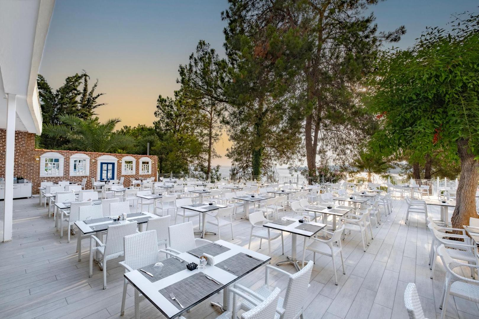 Utopia Beach Club plašā āra restorāna terase ar baltiem galdiem un krēsliem, ko ieskauj priedes un skats uz jūru