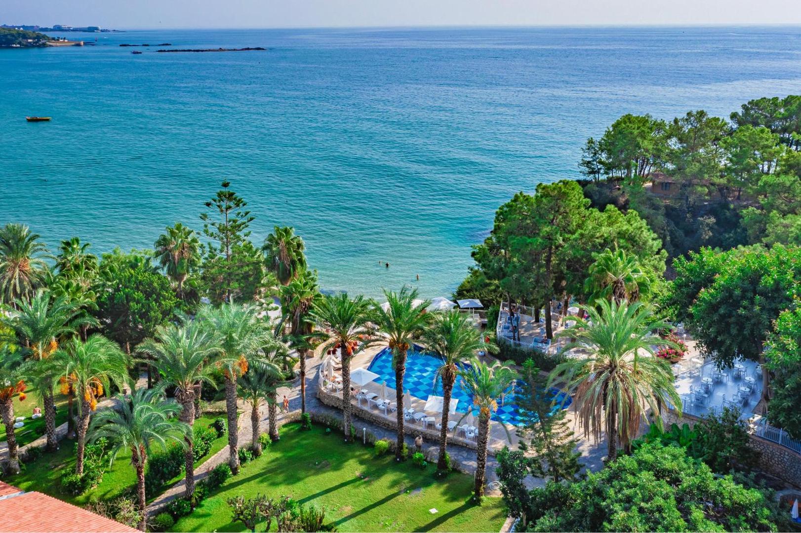 Utopia Beach Club panorāmas skats uz Vidusjūras līci, zaļo dārzu un baseinu Alānijā — tirkīza ūdens un priežu meža ieskauta teritorija