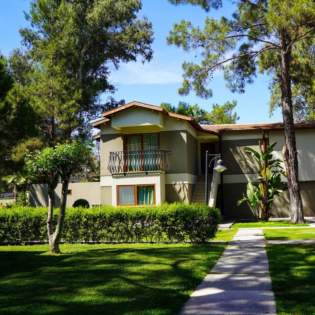 Kimeros Park Holiday Village divstāvu bungalo numurs ar balkonu, ko ieskauj kopts zālājs, dzīvžogs un augsti koki dārzā.