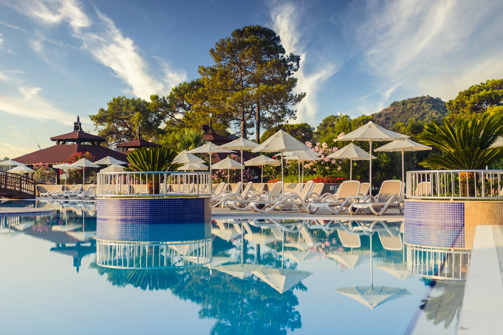 Kimeros Park Holiday Village atpūtas zona pie baseina ar sauļošanās krēsliem un saulessargiem — priedes un kalni fonā