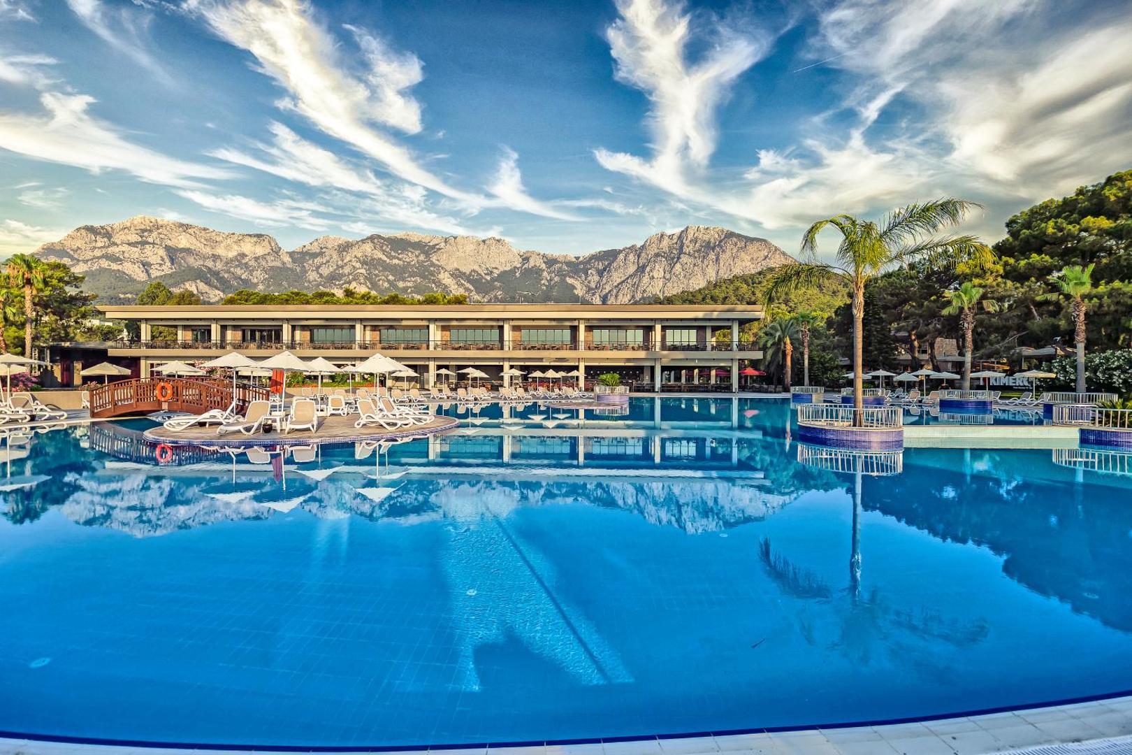 Kimeros Park Holiday Village plašais āra baseins ar skatu uz kalniem — sauļošanās krēsli, saulessargi un palmas piekrastē