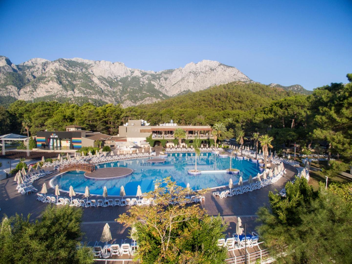 Kimeros Park Holiday Village galvenais āra baseins ar sauļošanās krēsliem un saulessargiem Kemeras reģionā, Turcijā, uz iespaidīgu kalnu fona.