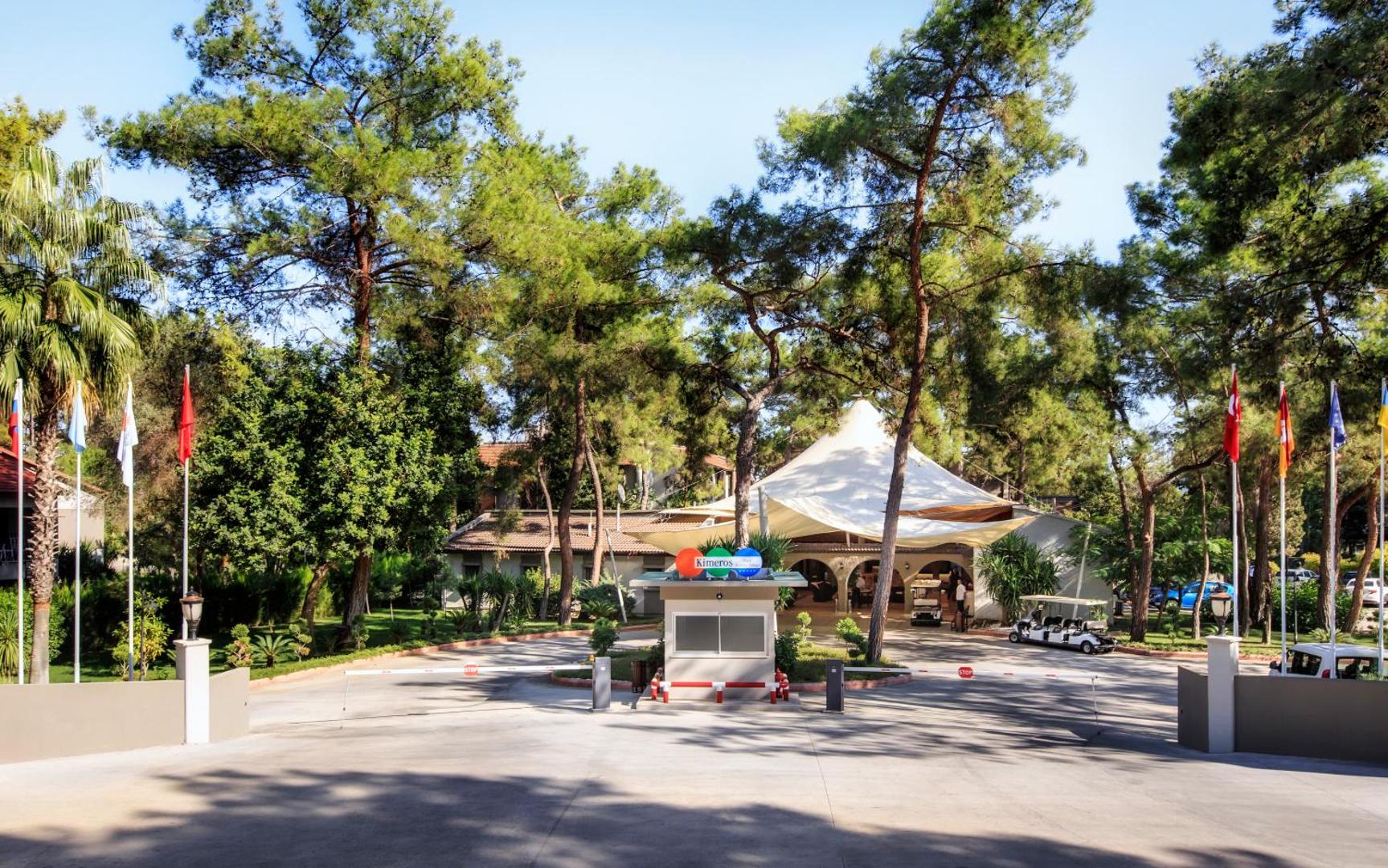 Kimeros Park Holiday Village galvenā ieeja ar karogiem, apsargājamu vārtu zonu un priežu kokiem viesnīcas teritorijā Turcijā.