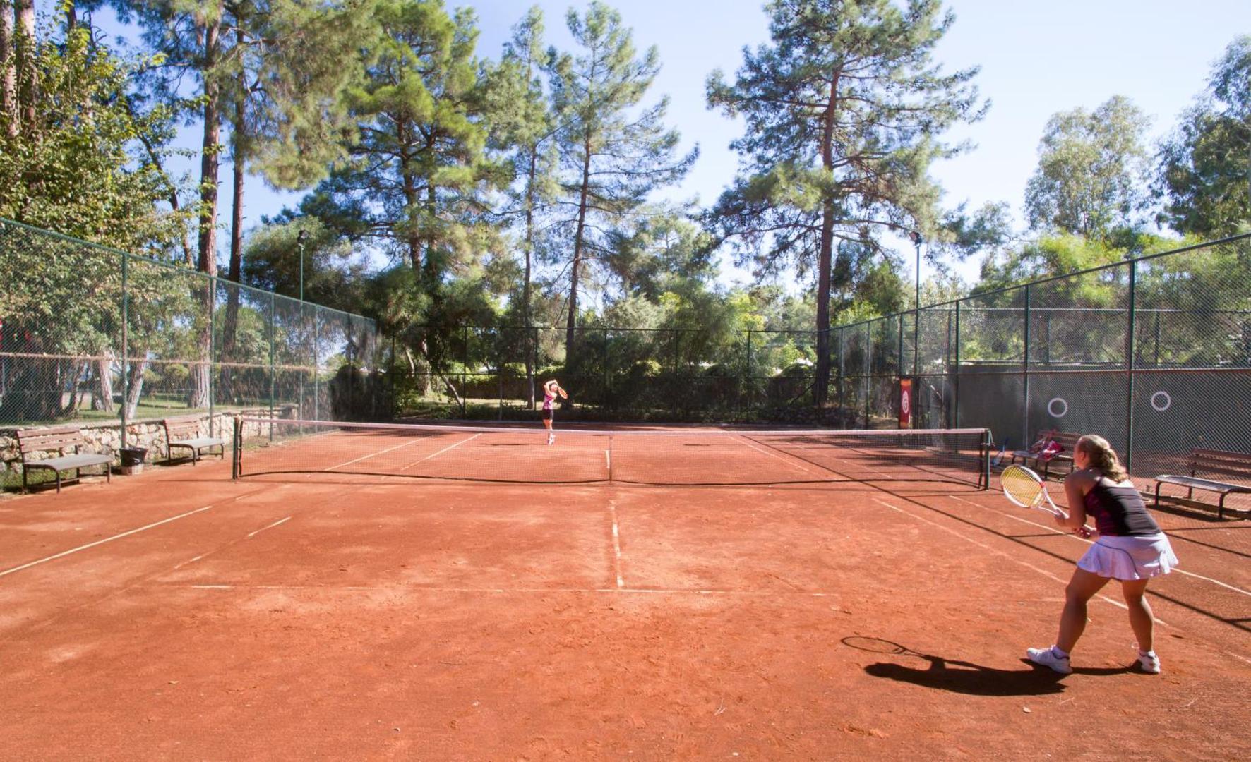 Kimeros Park Holiday Village tenisa korti ar māla segumu, ko ieskauj augstas priedes un zaļojoša daba Kemeras piekrastē