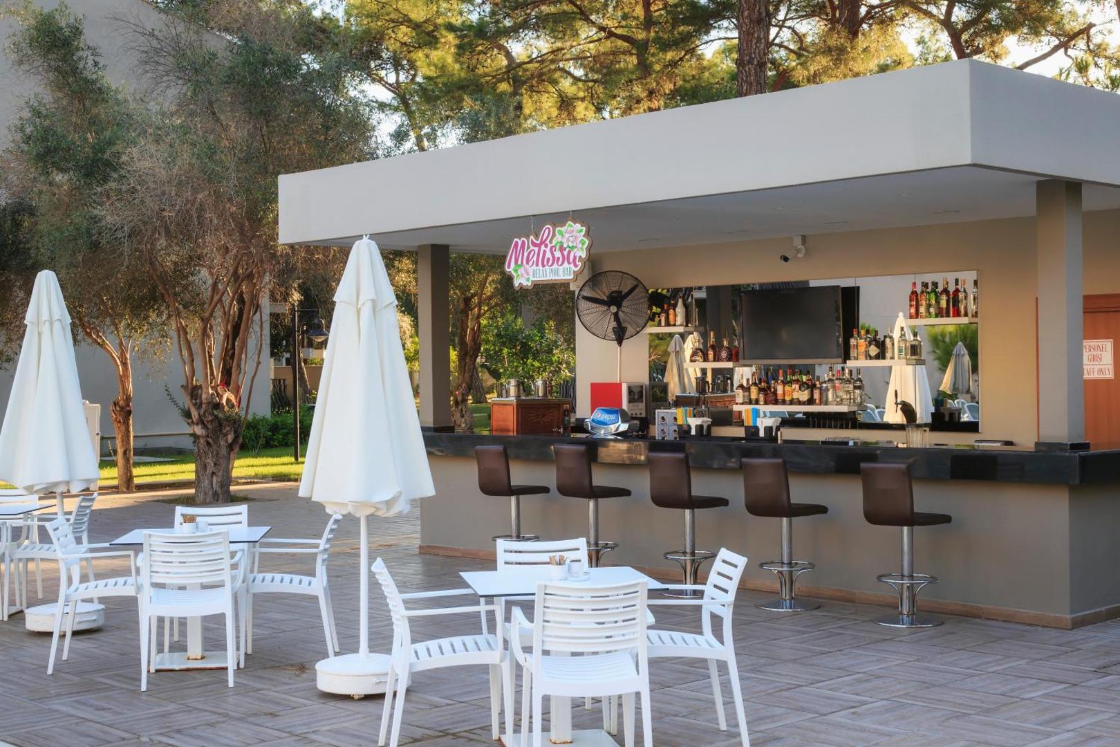 Kimeros Park Holiday Village āra bārs Melissa priežu meža ielokā ar baltiem galdiņiem un saulessargiem Kemeras kūrortā