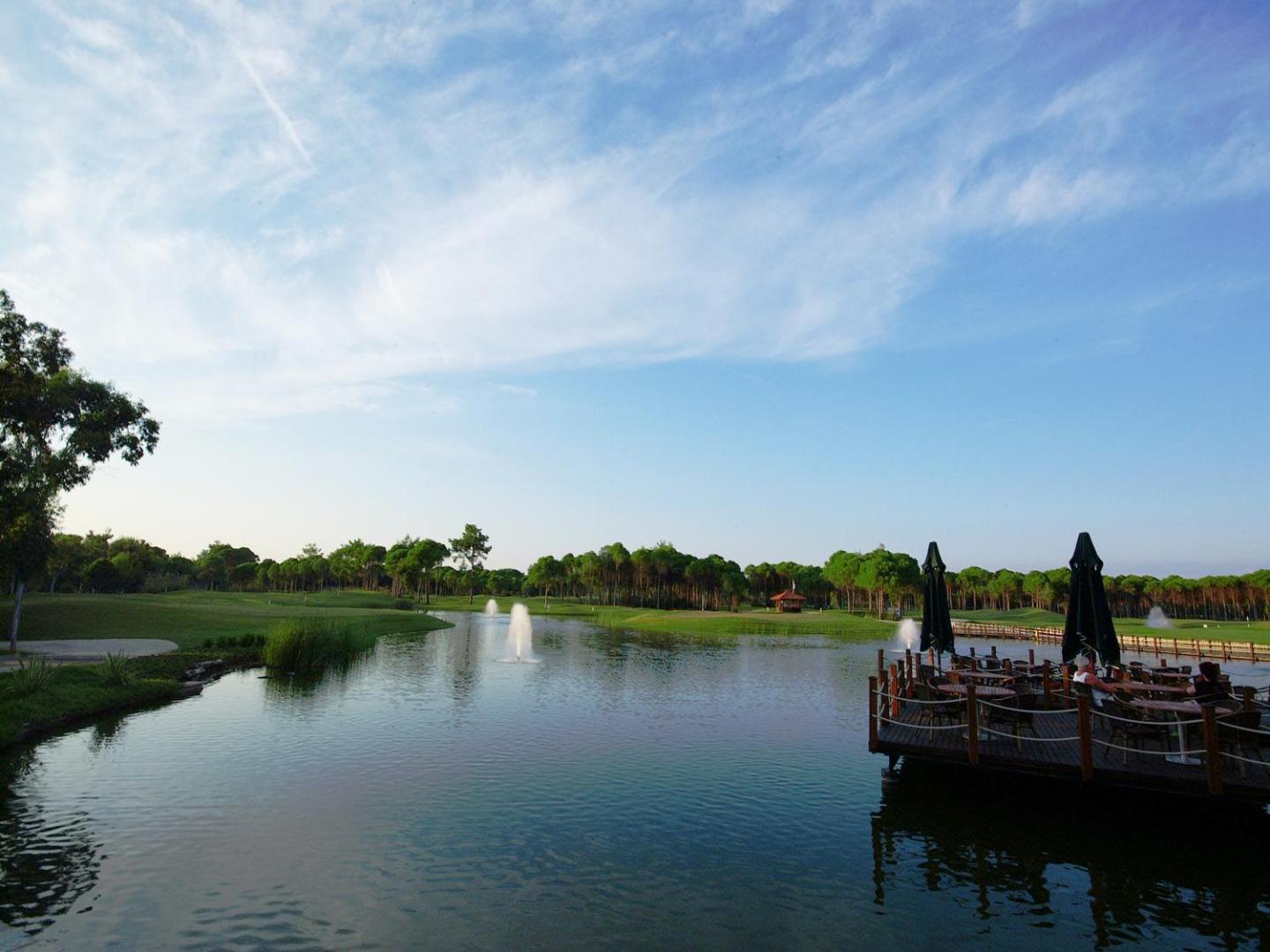 Sueno Hotels Deluxe Belek golfa laukums ar ezeru un strūklakām — skats no koka terases uz zaļo zonu un mežu