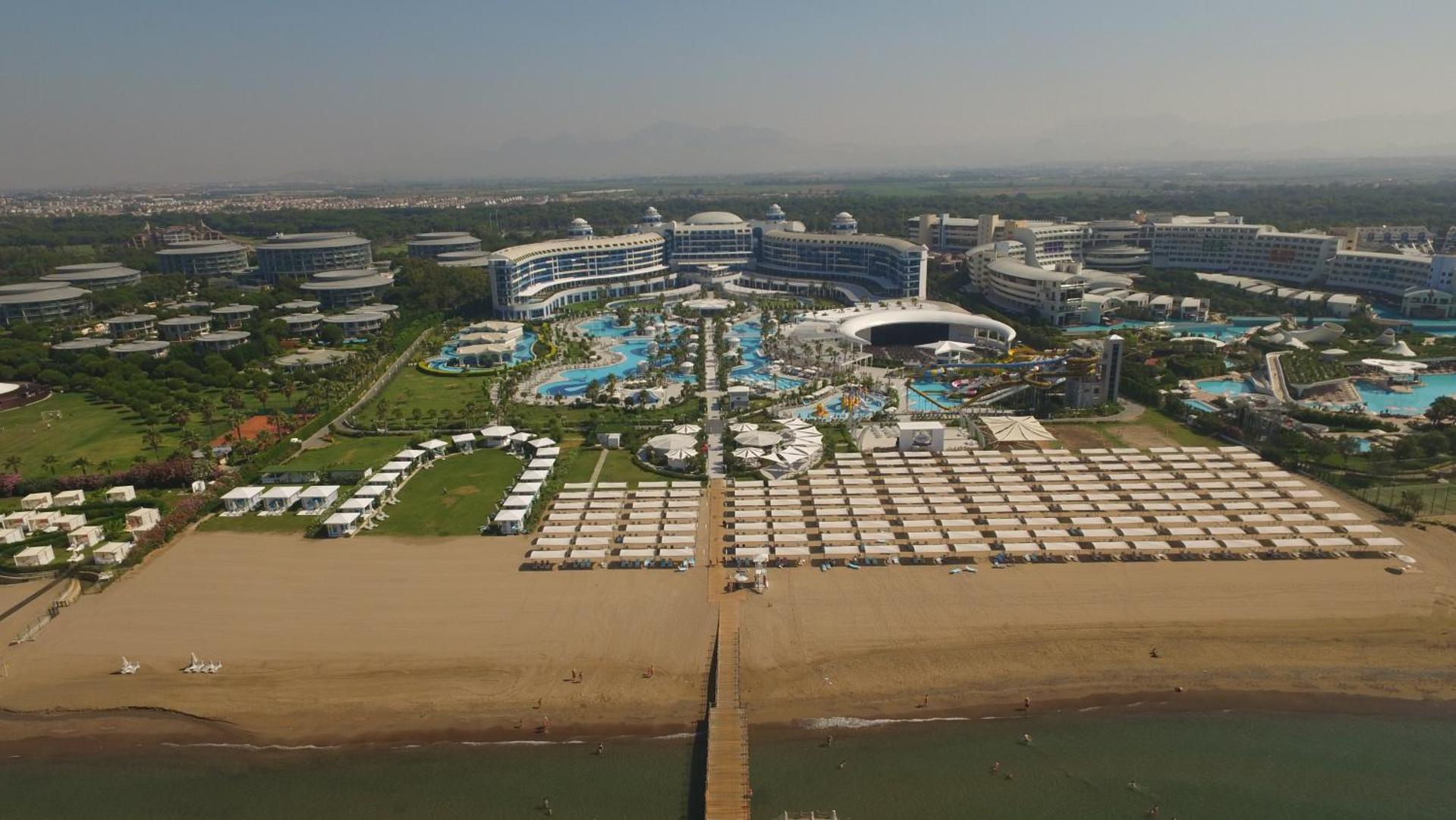 Sueno Hotels Deluxe Belek panorāmas skats uz privāto pludmali, saulessargu zonu un viesnīcas galveno ēku Belekā, Turcijā