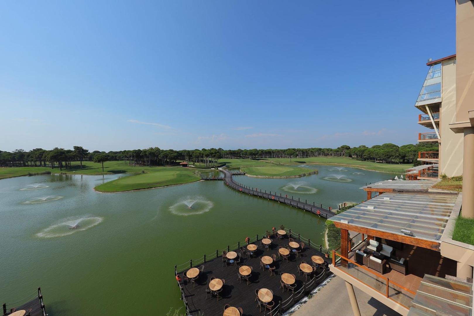Sueno Hotels Golf Belek āra terase ar galdiņiem virs ūdens un skatu uz golfa laukumiem un strūklakām ezerā