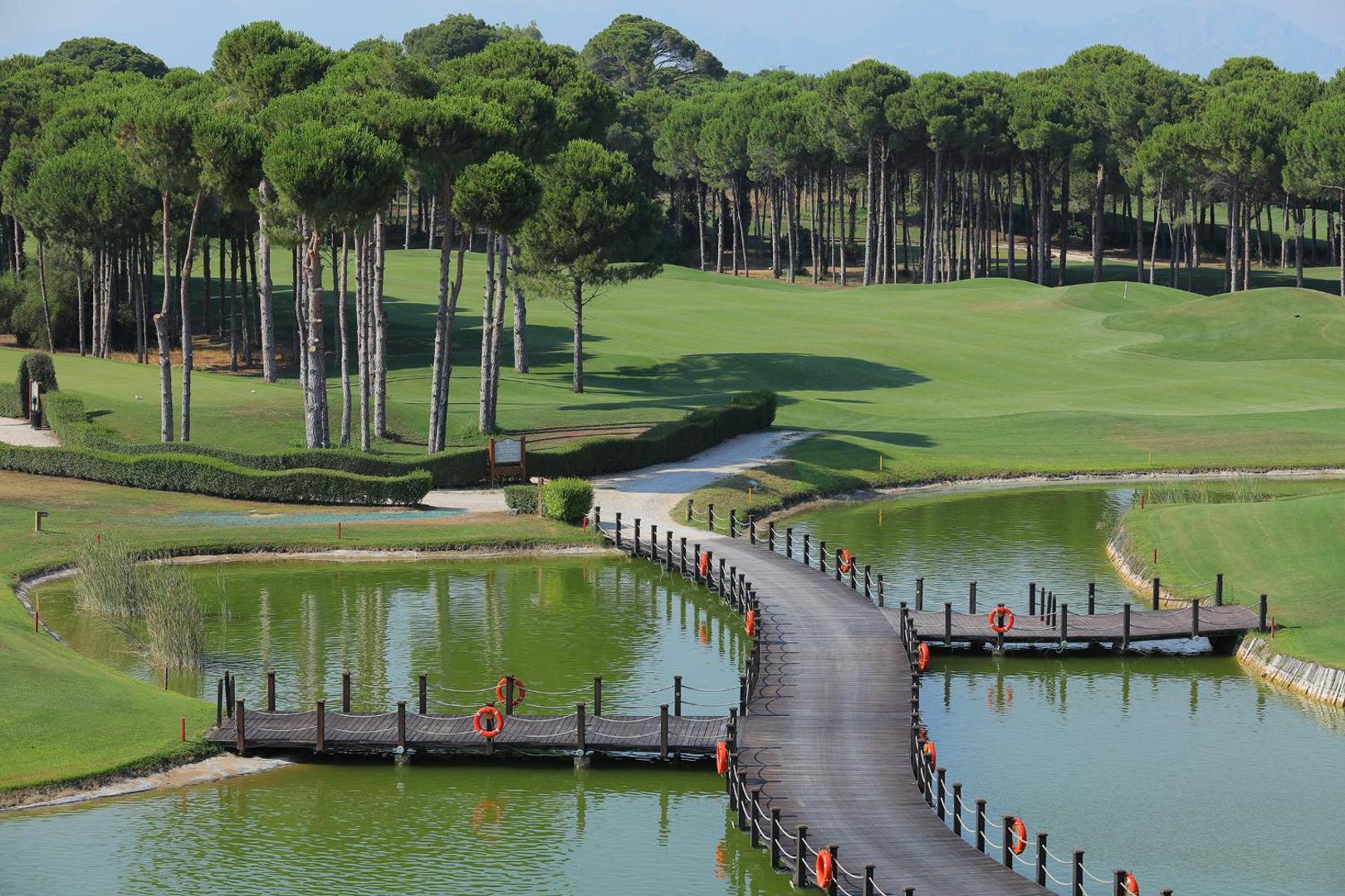 Sueno Hotels Golf Belek golfa laukums ar koka tiltiņu pār dīķi, priežu mežu un koptu zālienu Belekas reģionā