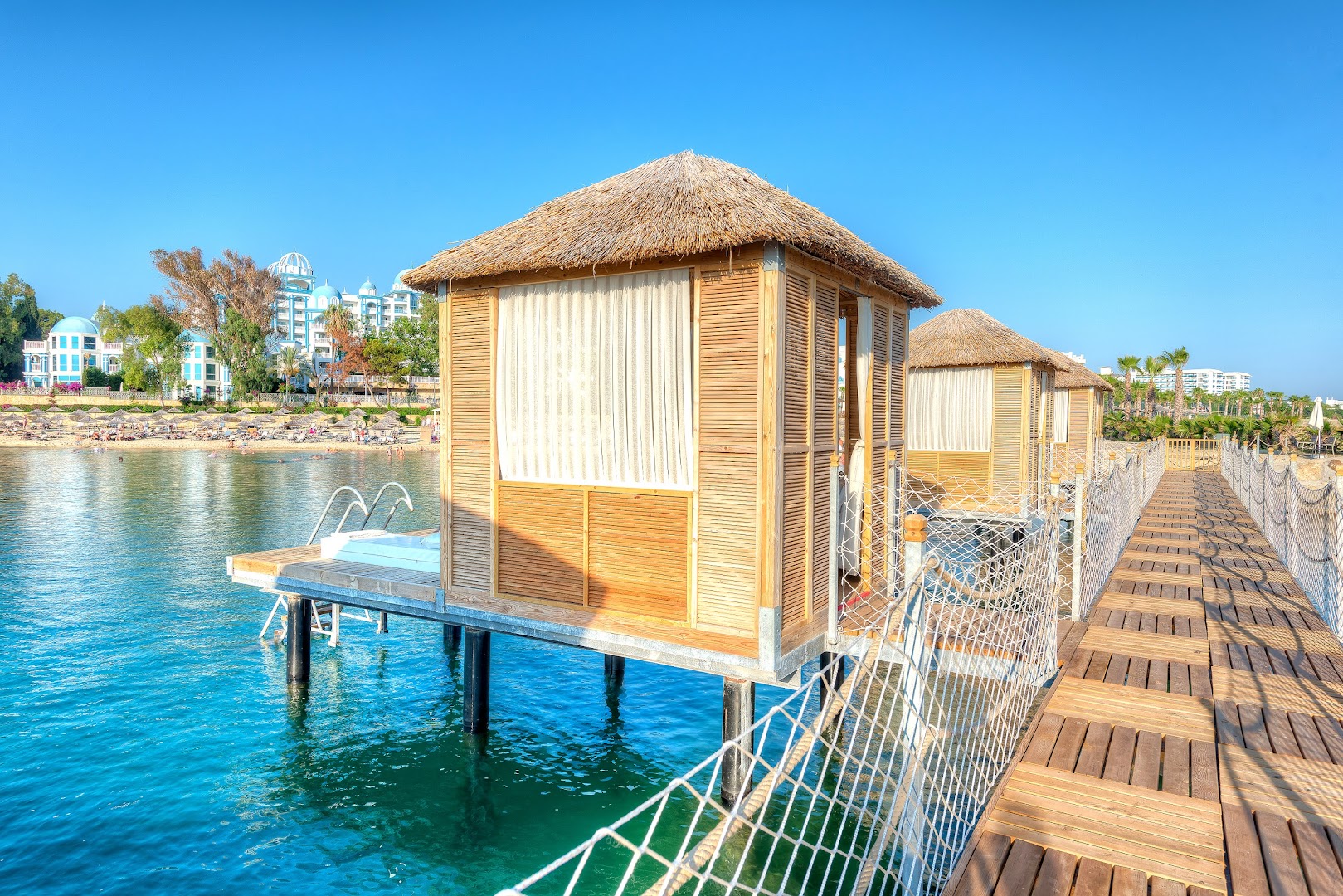 Rubi Platinum SPA Resort & Suites koka bungalo uz pāļiem virs ūdens ar kāpnēm jūrā un viesnīcas ēku fonā Alānijā