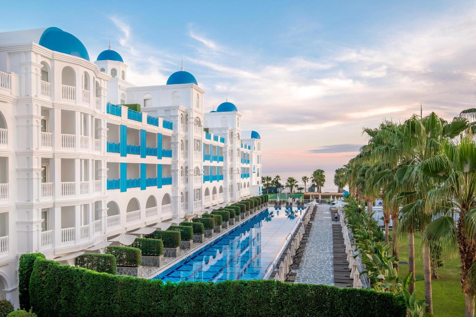 Rubi Platinum SPA Resort & Suites modernā arhitektūra ar zilajiem kupoliem un garu peldbaseinu saulrieta gaismā Alanjas piekrastē