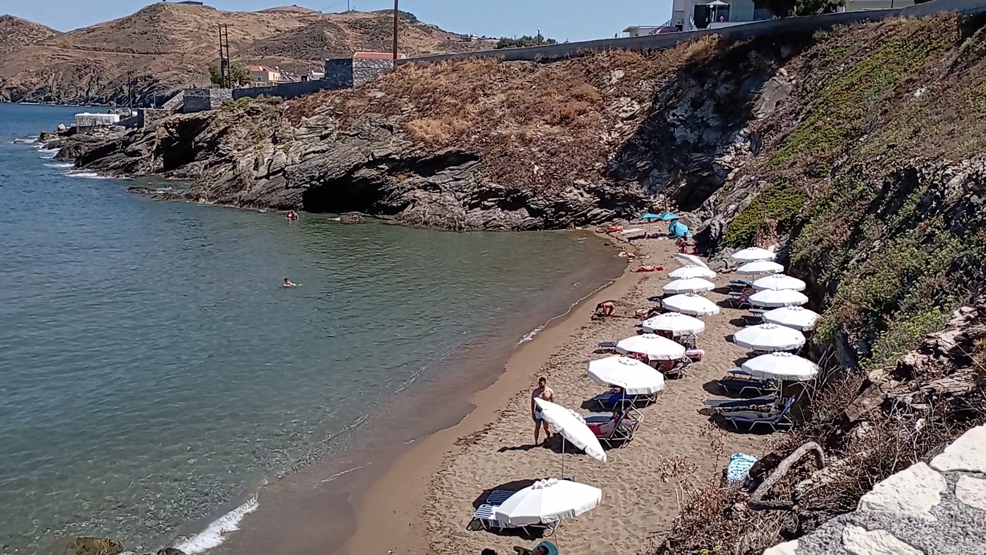 Panormo Beach mājīgā pludmales zona klinšu ielokā — balti saulessargi un sauļošanās krēsli pie mierīga, caurspīdīga ūdens