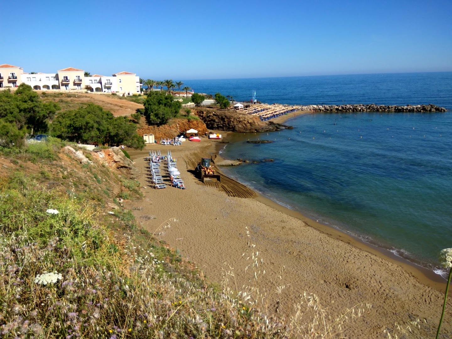 Panormo Beach viesnīcas panorāmas skats uz smilšu pludmali un zilo jūru Krētā — piekrastes zona ar saulessargiem un viesnīcas ēkām fonā
