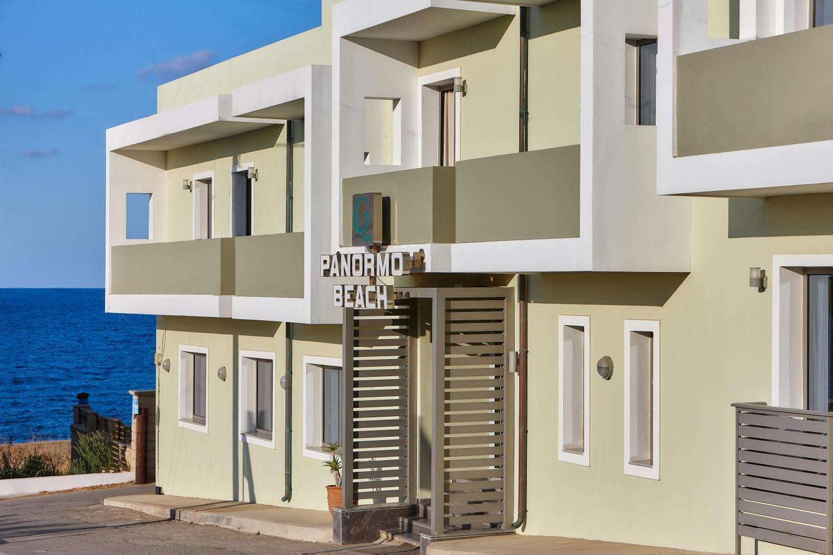 Panormo Beach viesnīcas fasāde Krētā ar skatu uz jūru — gaiša ēka ar balkoniem un ieeju piekrastes ciematā