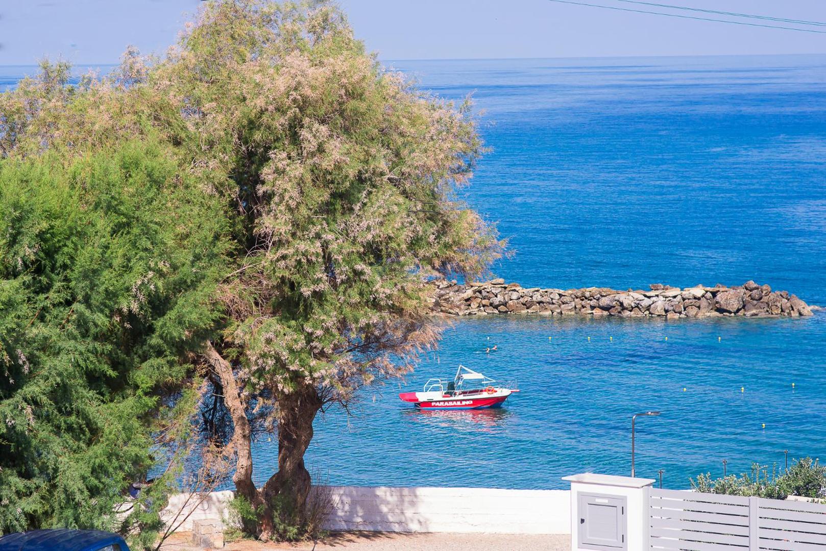 Panormo Beach viesnīcas skats uz zilo jūru caur zaļojošiem kokiem, kur līcī noenkurota sarkana motorlaiva Panormo ciemā, Krētā