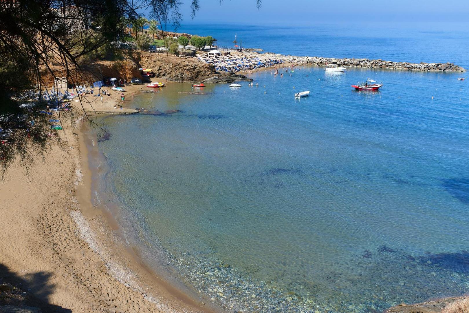 Panormo ciemata pludmale ar smiltīm, saulessargiem un nelielām laivām dzidrā līča ūdenī netālu no Panormo Beach viesnīcas