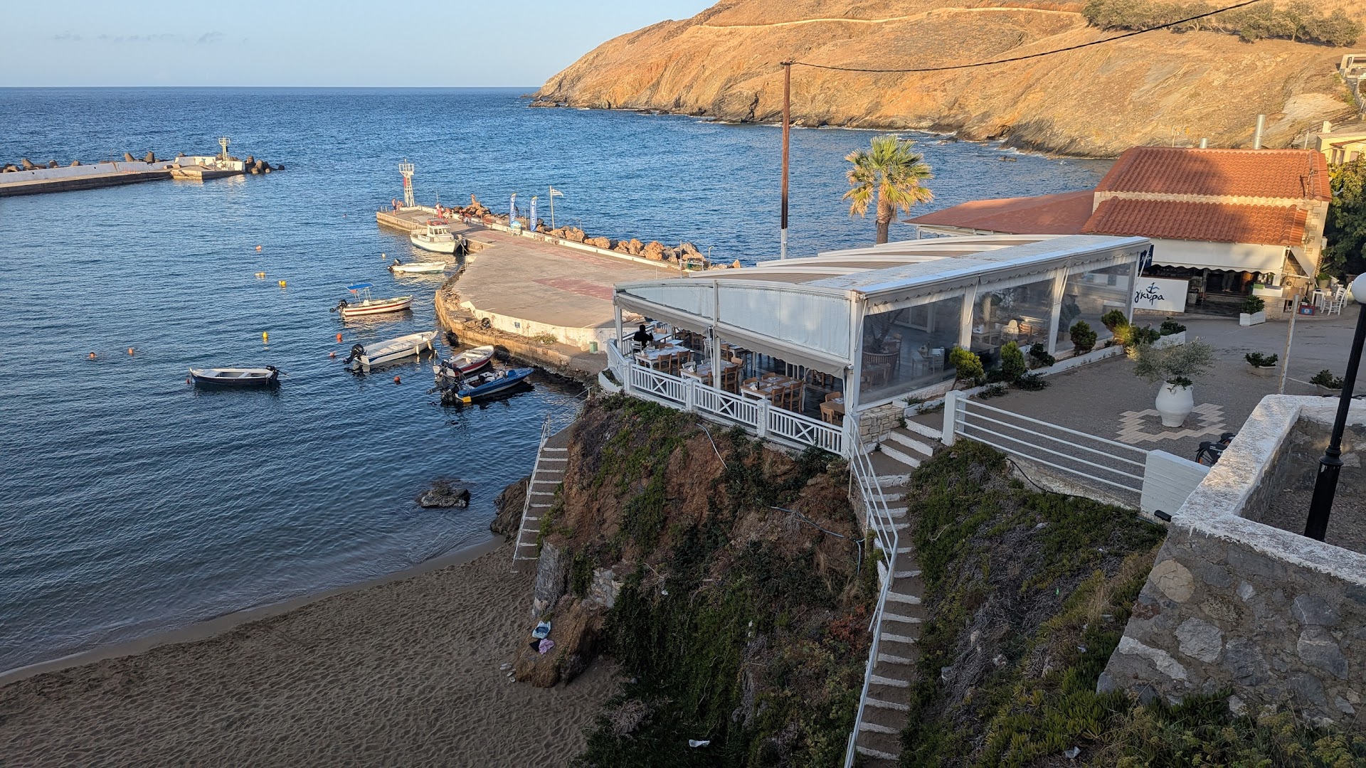 Panormo Beach āra terase uz klints malas ar skatu uz Panormo ostu, laivām un smilšu pludmali saulrietā
