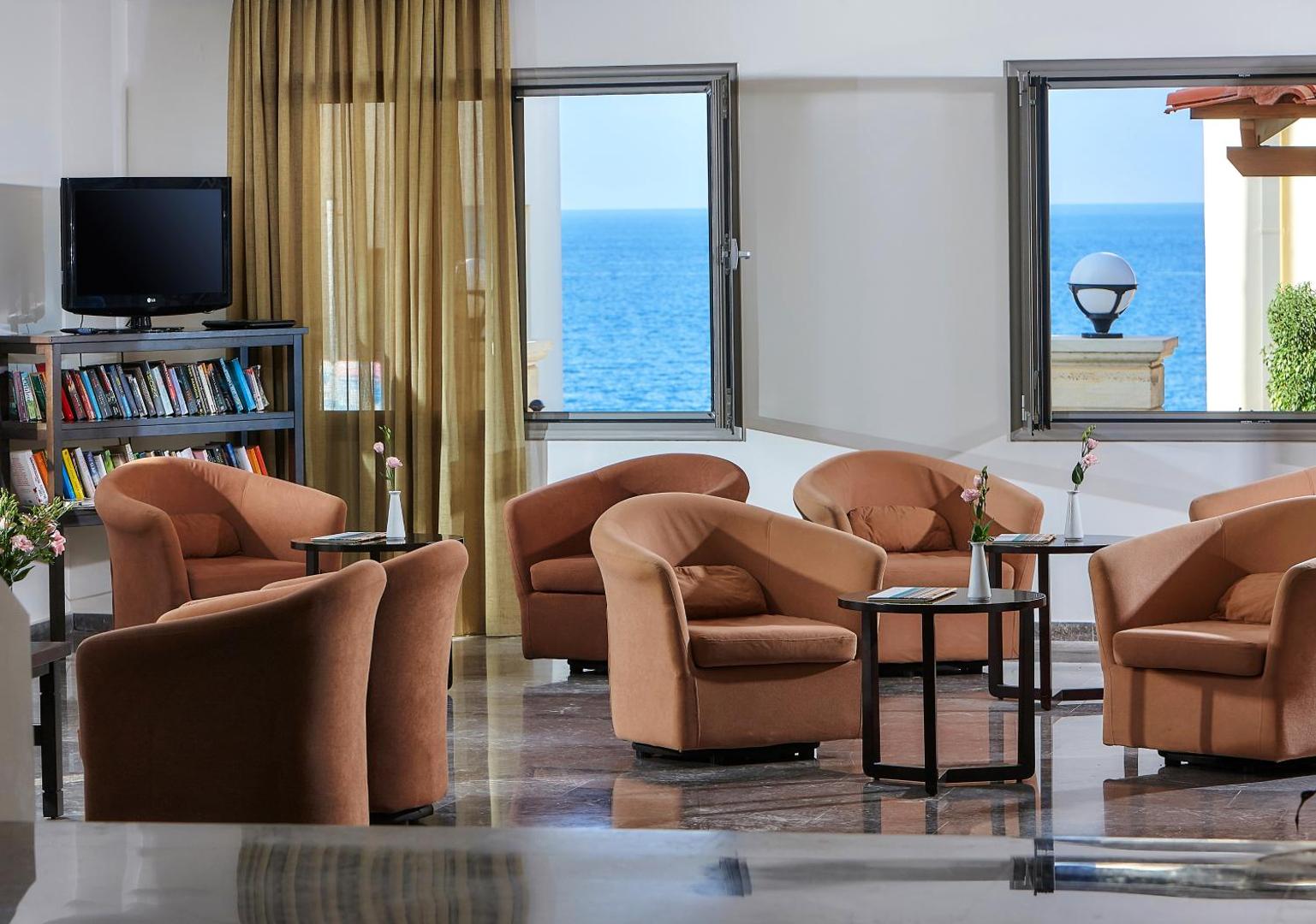 Panormo Beach viesnīcas lobby atpūtas zona ar ērtiem krēsliem, bibliotēku un lieliem logiem, no kuriem paveras skats uz jūru