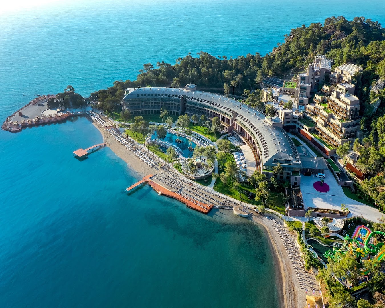 NG Phaselis Bay viesnīcas teritorija no augšas — skats uz galveno ēku, pludmales līniju un akvaparku bērniem Kemeras piekrastē