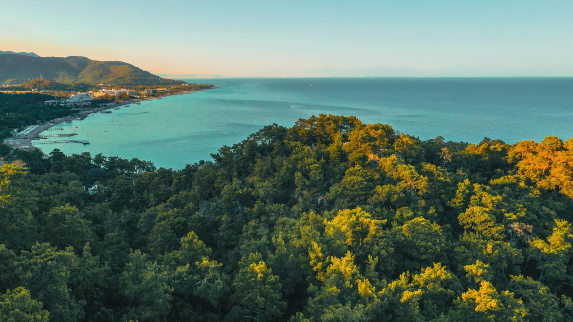 NG Phaselis Bay viesnīcas apkārtne no augšas — blīvs priežu mežs piekrastē un skats uz līci Kemeras reģionā saulrieta gaismā