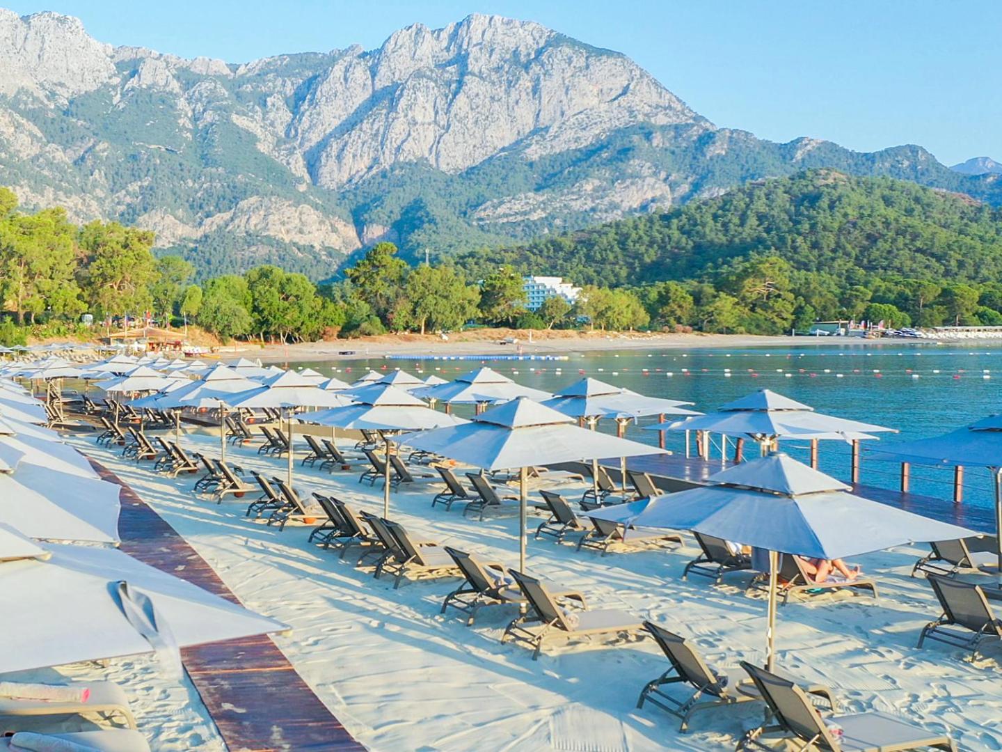 NG Phaselis Bay privātā smilšu pludmale ar saulessargiem un sauļošanās krēsliem uz kalnu fona Kemeras reģionā, Turcijā