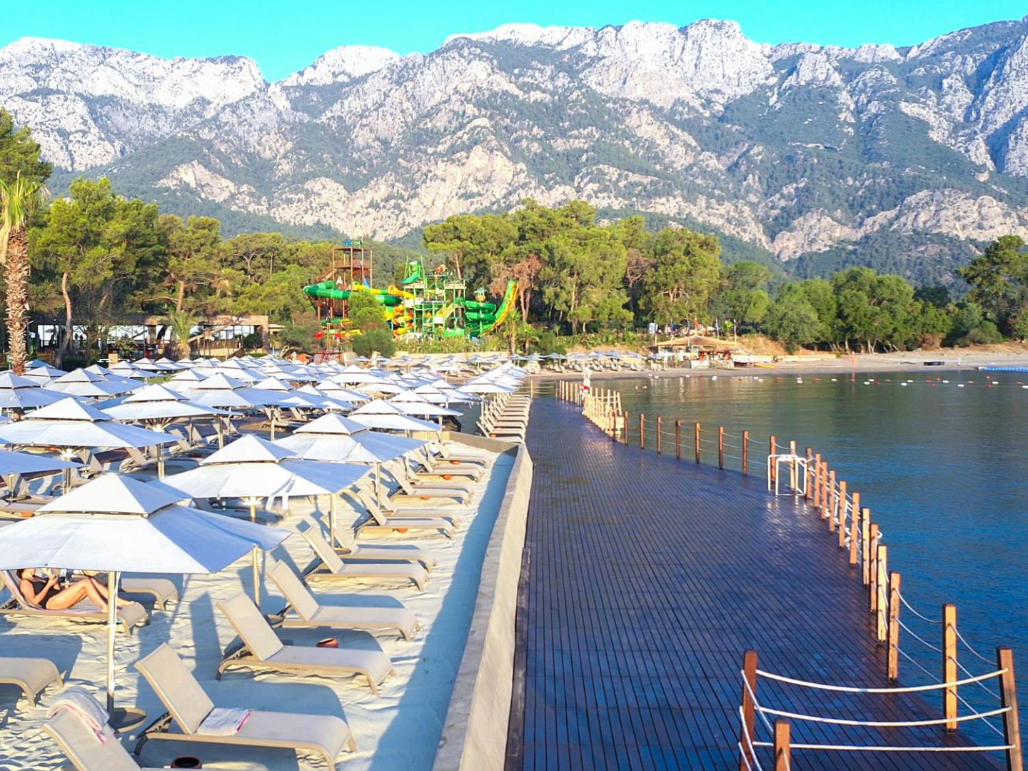 NG Phaselis Bay pludmales zona ar koka laipu un skatu uz akvaparku un kalniem Kemeras piekrastē