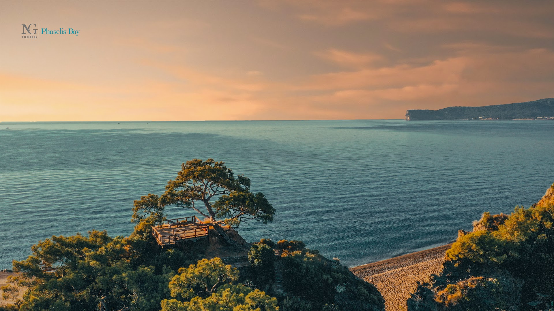 NG Phaselis Bay skatu vieta uz klints saulrieta laikā — koka terase zem priedes ar skatu uz jūru un pludmali siltos toņos