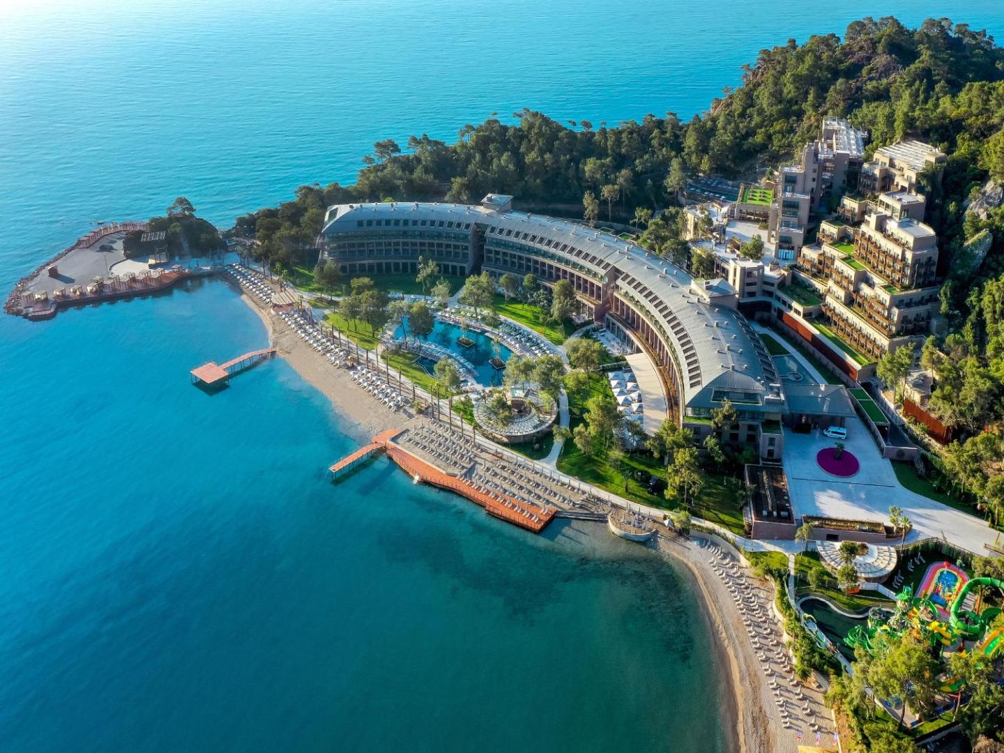 NG Phaselis Bay viesnīcas panorāmas skats no putna lidojuma Kemeras reģionā — privātais līcis, smilšu pludmale un baseinu zona priežu meža ieskāvumā