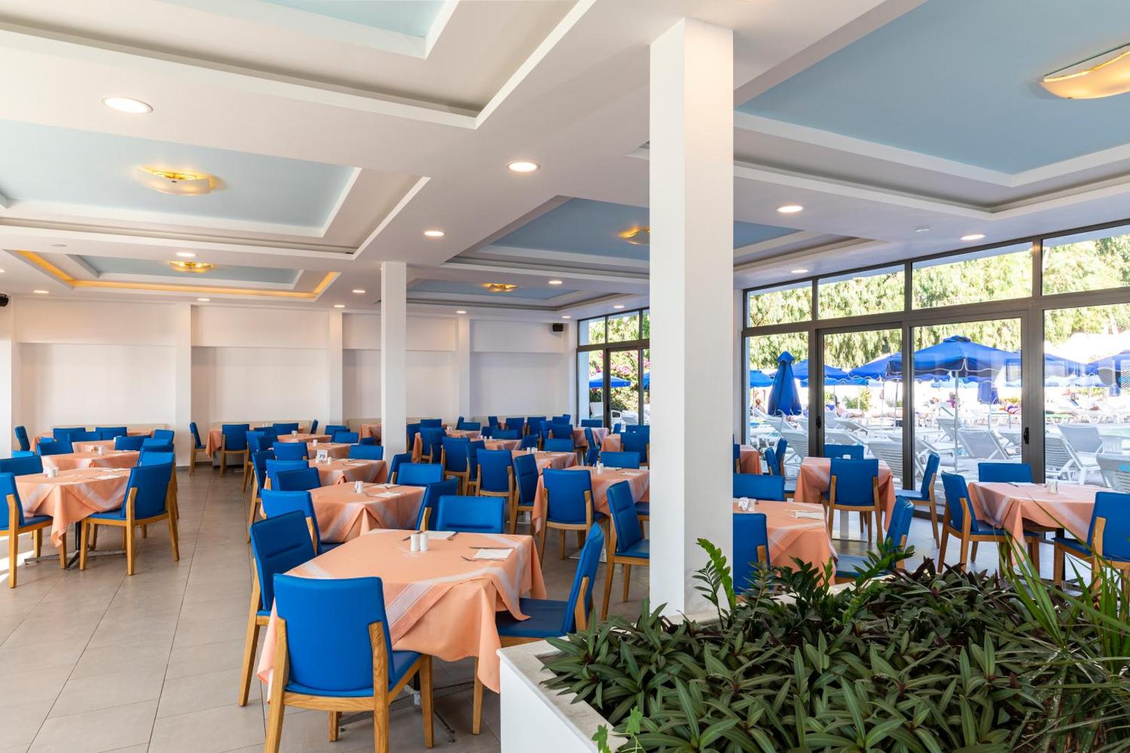 Belair Beach galvenais restorāns ar klātiem galdiem un zilām mēbelēm — plaši logi ar skatu uz baseina zonu