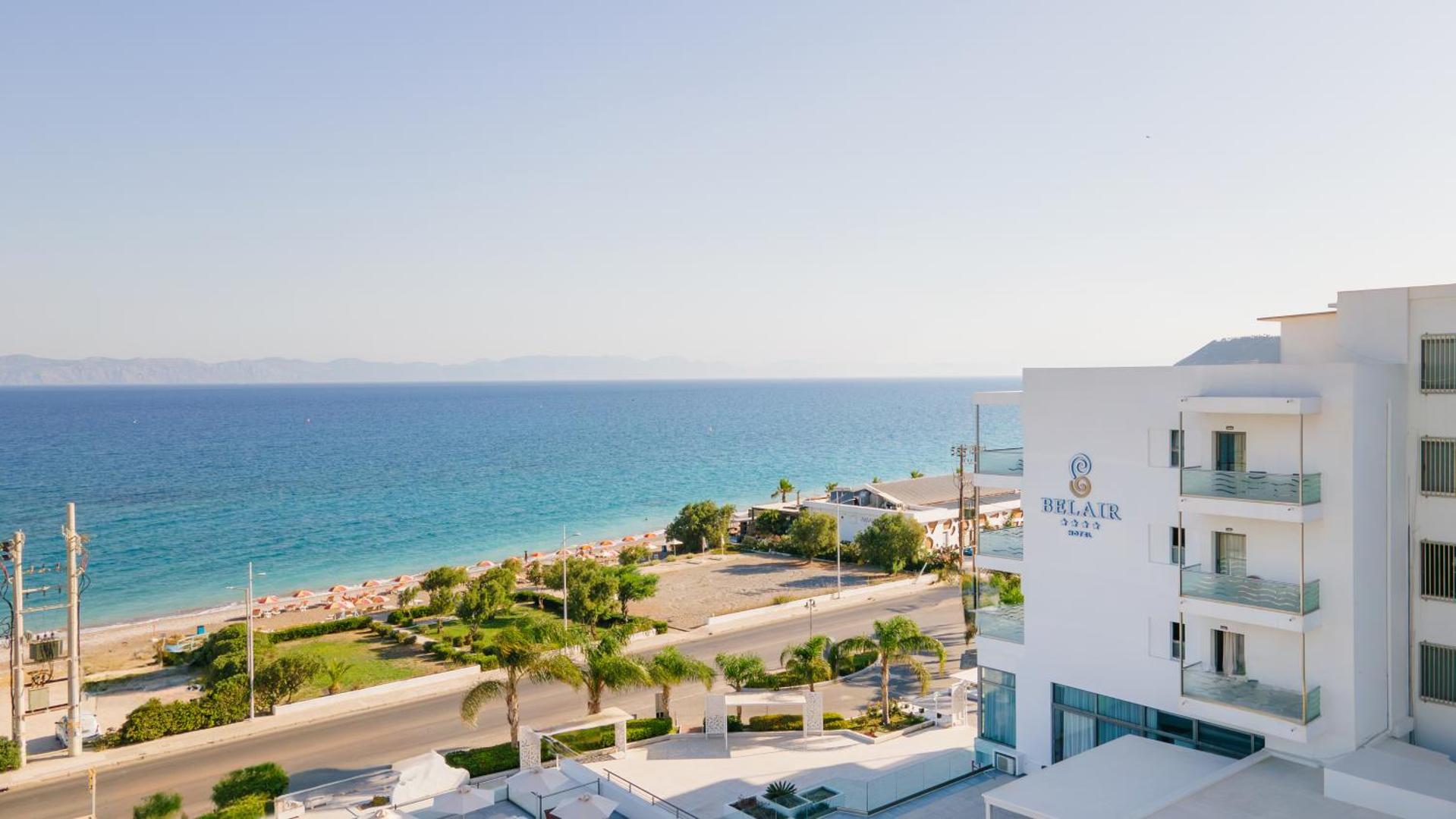 Belair Beach viesnīcas fasāde ar logo un skatu uz pludmali pāri ceļam Rodā — palmas un zilais jūras horizonts