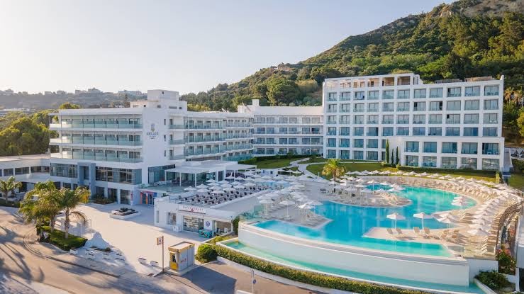 Belair Beach viesnīcas galvenais baseins ar saulessargiem un zviļņiem Rodā — moderna arhitektūra un kalnu fons saulainā dienā