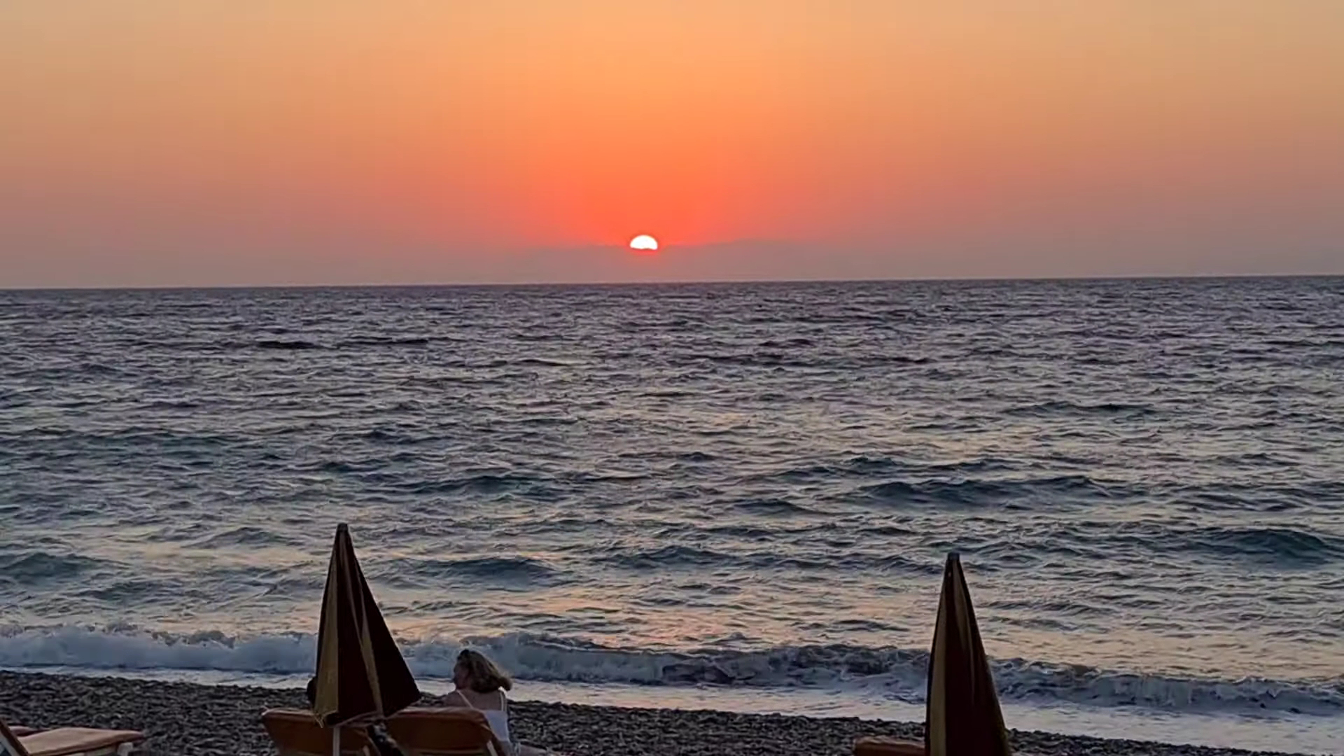 Saulriets virs Egejas jūras Belair Beach viesnīcas pludmalē Rodā — oļu krasts ar saulessargiem un oranžām debesīm