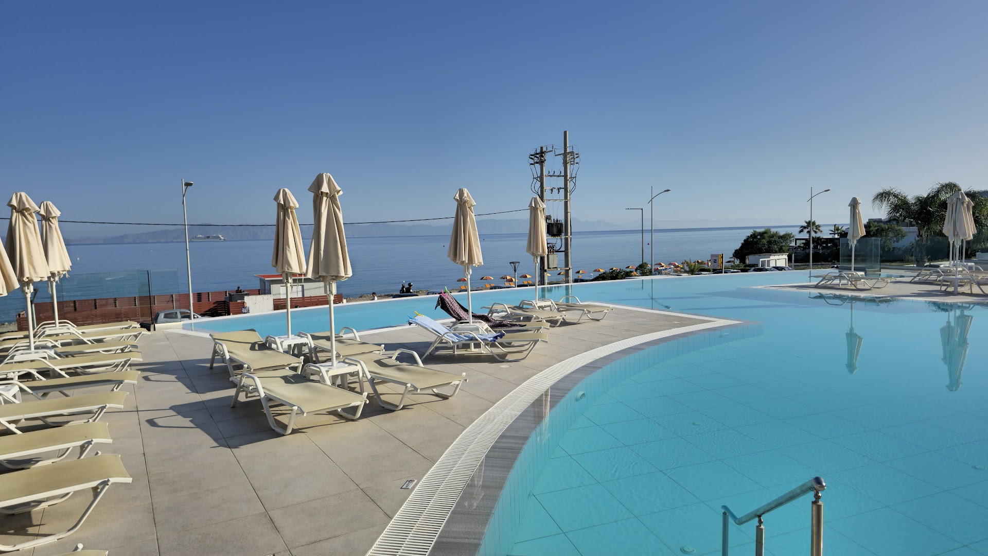 Belair Beach baseina zona ar atpūtas krēsliem un skatu uz jūru un kalniem tālumā Rodas salas piekrastē