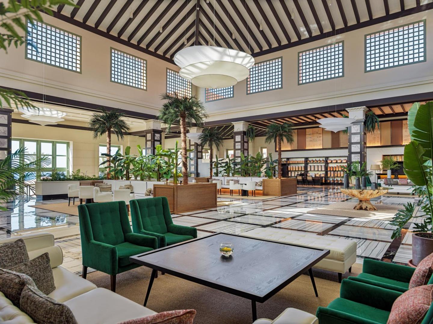 H10 Costa Adeje Palace viesnīcas plašais vestibils Tenerifē ar augstiem griestiem, palmām un stilīgiem zaļiem atpūtas krēsliem gaišā interjerā