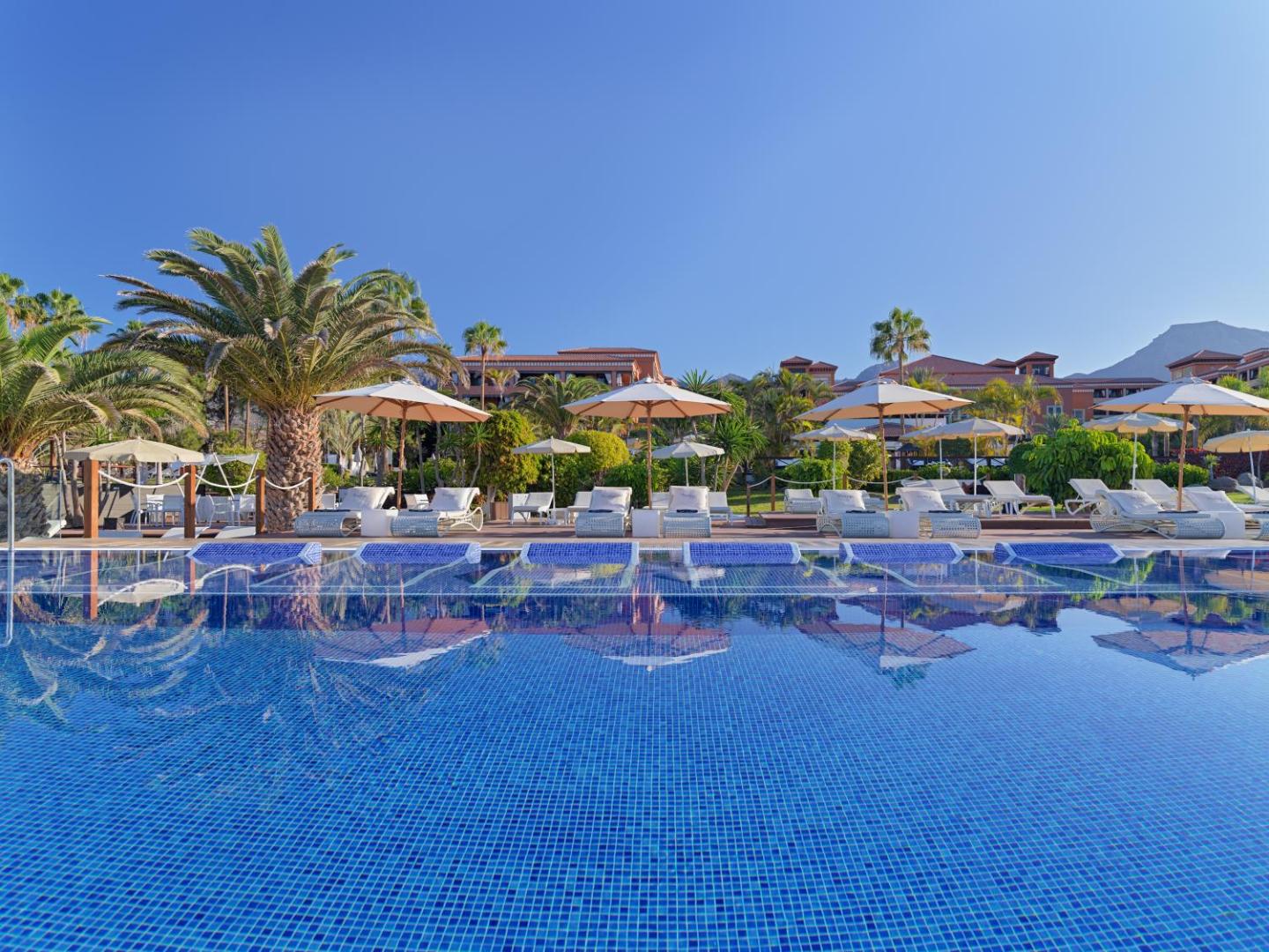 H10 Costa Adeje Palace āra baseins ar sauļošanās zonu, palmām un viesnīcas ēku fonā zem skaidrām debesīm