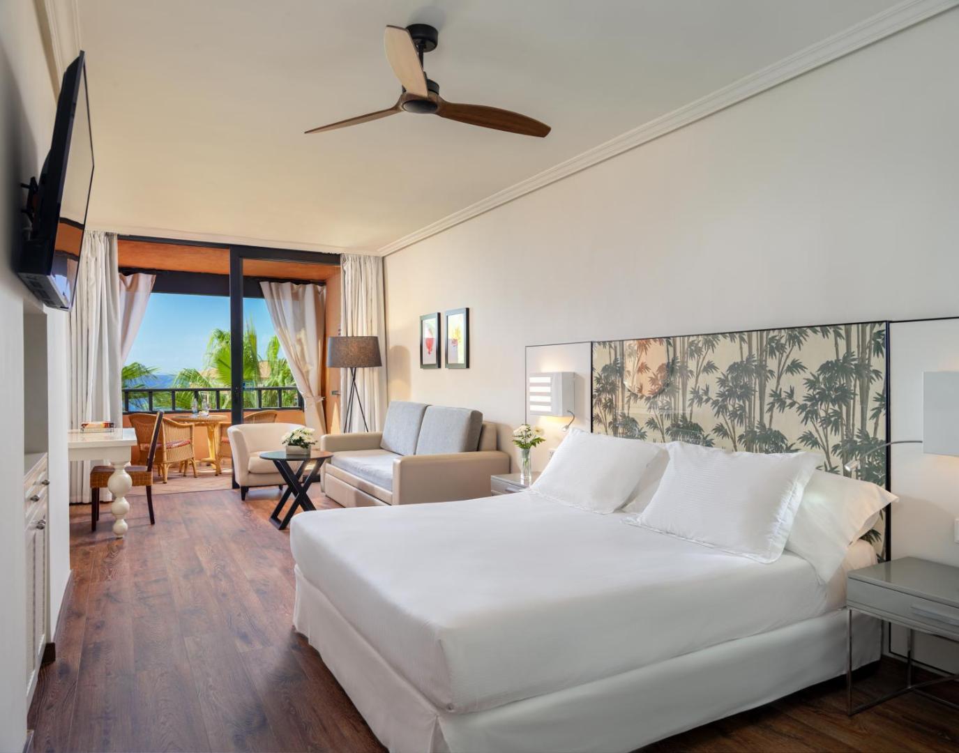 Viesnīcas H10 Costa Adeje Palace numurs ar dīvānu, lielu gultu un balkonu, no kura paveras skats uz palmām un okeānu