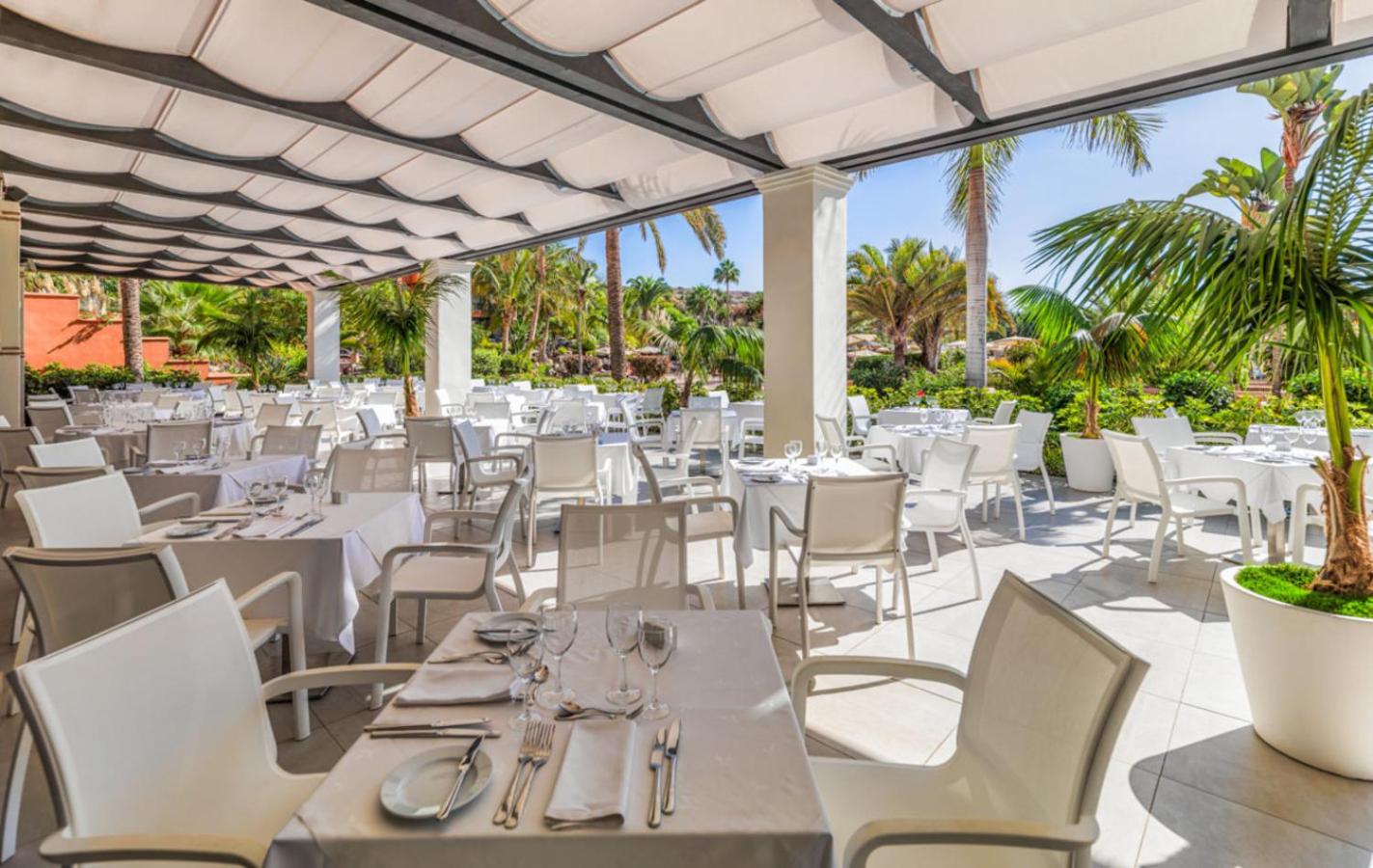 H10 Costa Adeje Palace āra terases restorāns ar baltiem galdiem zem nojumes, ko ieskauj palmas un tropu dārzs.