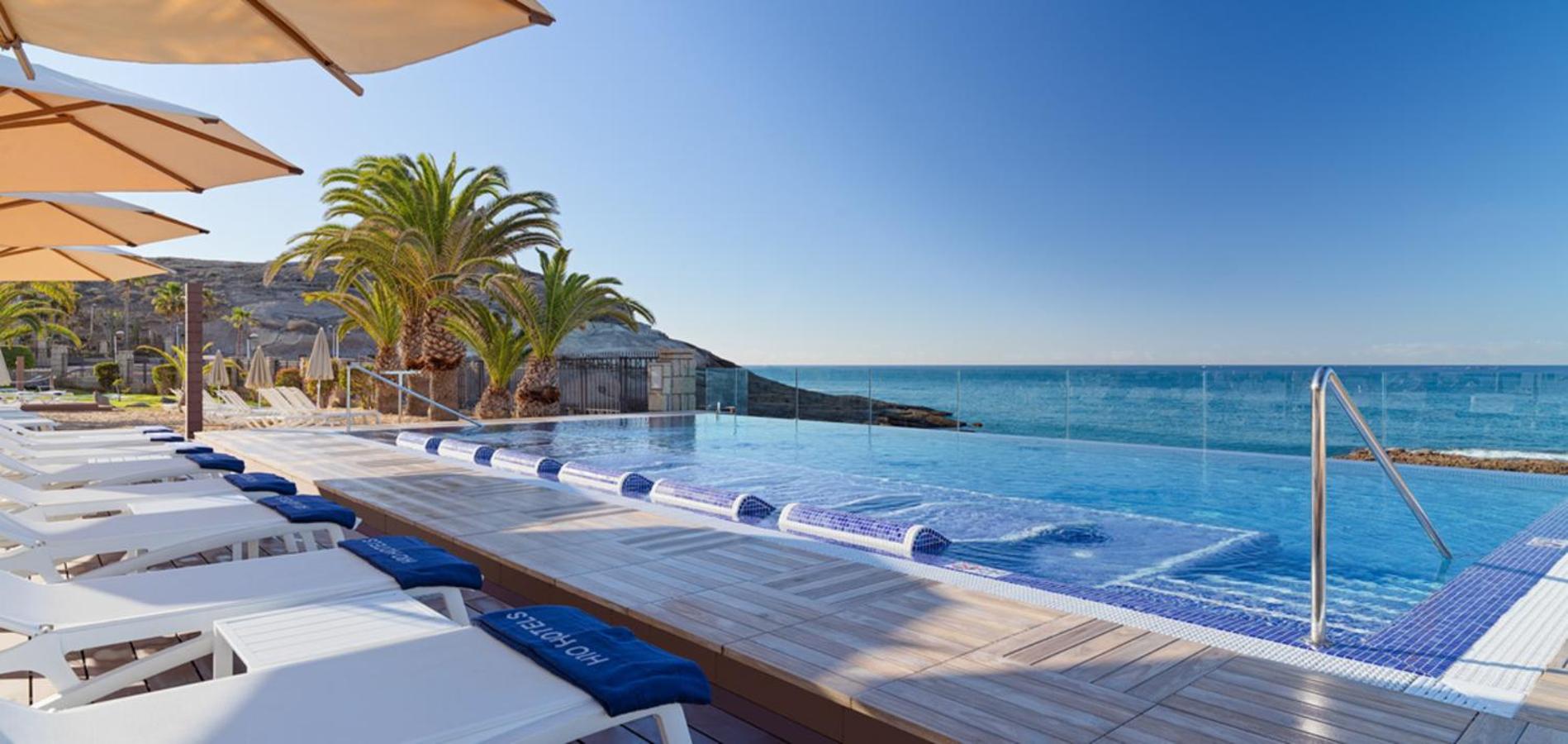 H10 Costa Adeje Palace bezgalības baseins ar skatu uz okeānu, sauļošanās krēsliem un saulessargiem piekrastē Tenerifē