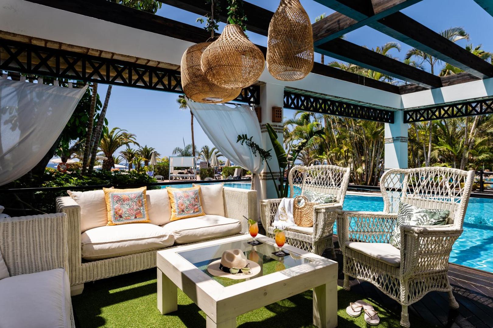 H10 Costa Adeje Palace āra terase ar pītiem dīvāniem un krēsliem pie baseina, ko ieskauj palmas un tropu dārzs