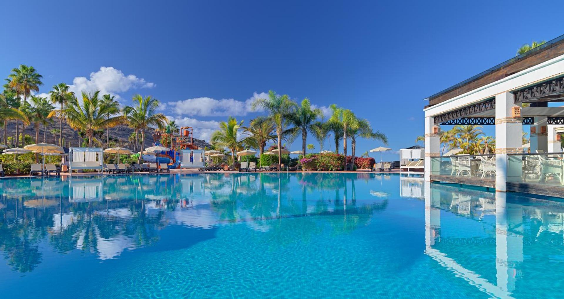 H10 Costa Adeje Palace plašais āra peldbaseins Tenerifē ar palmām, saulessargiem un bērnu ūdens atrakciju parku fonā zem skaidrām debesīm.