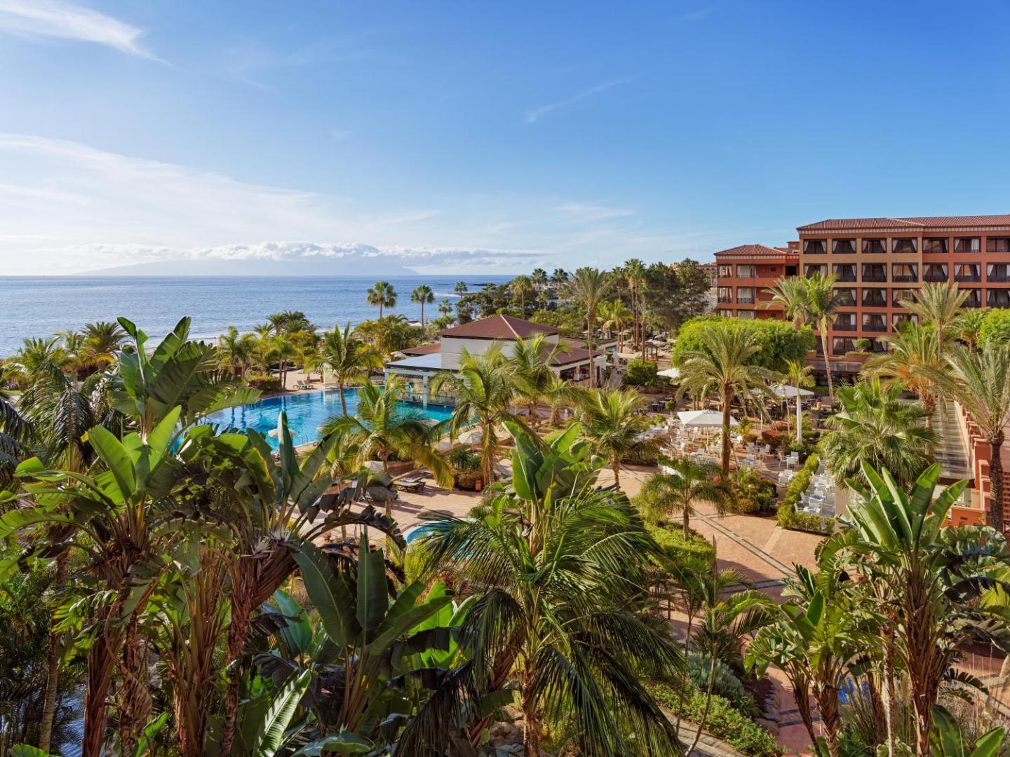 H10 Costa Adeje Palace panorāmas skats uz baseinu zonu, palmu dārzu un okeāna piekrasti Tenerifē, Kanāriju salās saulainā dienā