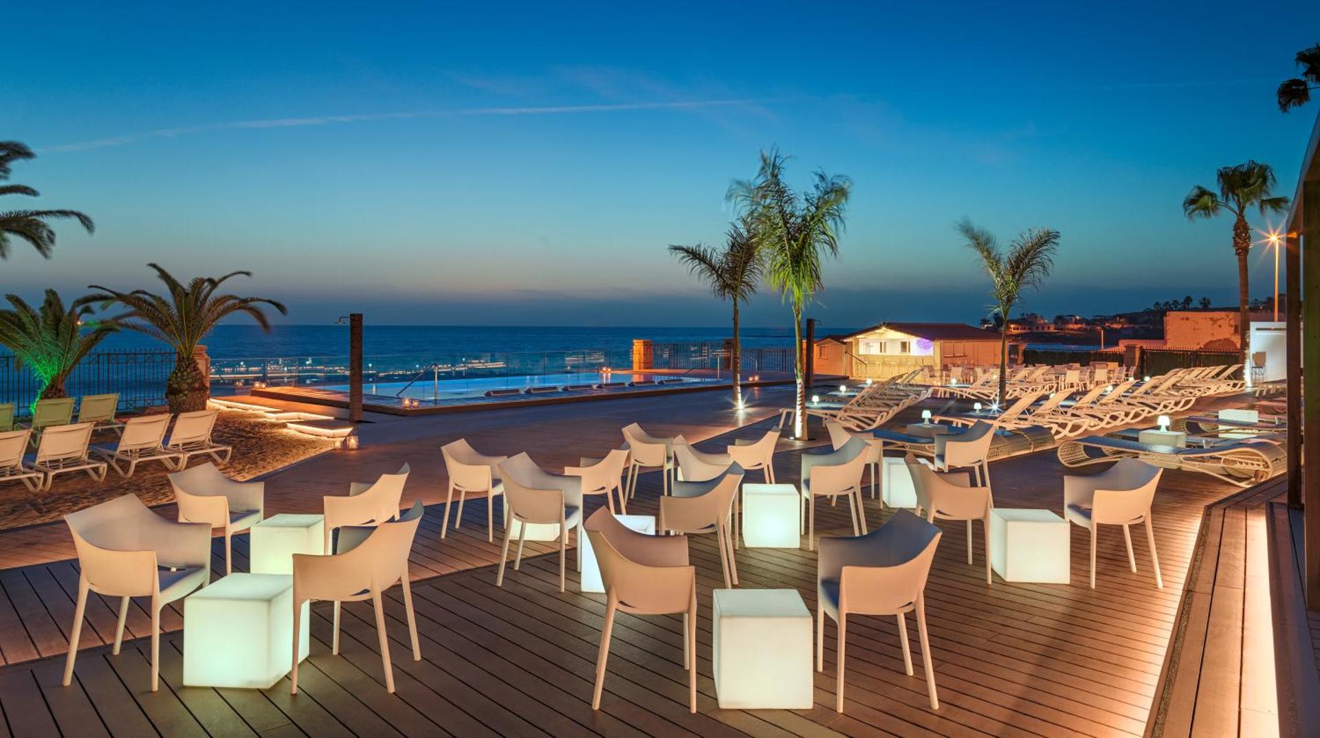 H10 Costa Adeje Palace vakara skats uz āra terasi ar izgaismotām mēbelēm, baseinu un okeāna horizontu saulrietā