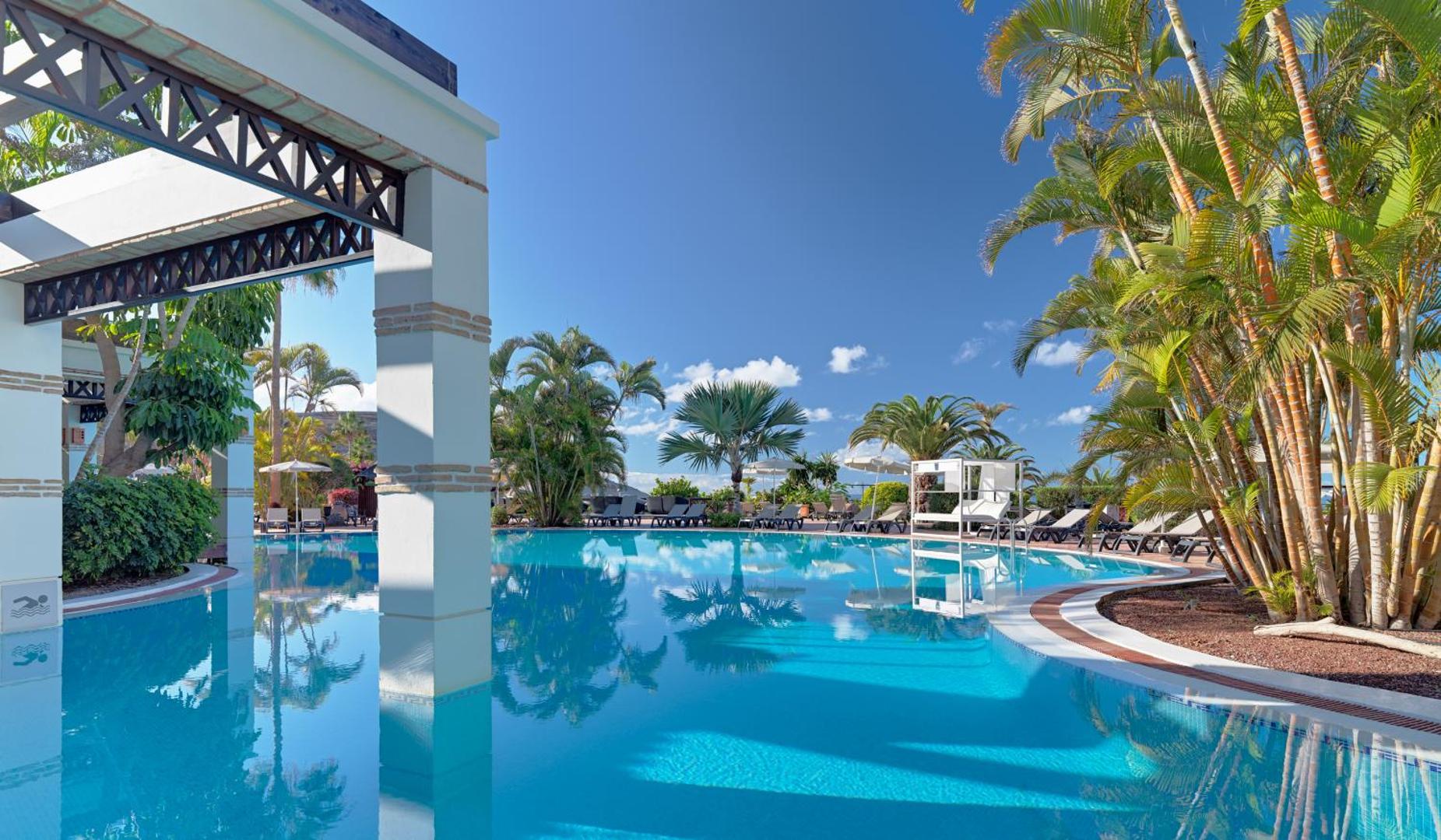 H10 Costa Adeje Palace viesnīcas dārza baseins ar palmām un atpūtas krēsliem, atspulgi ūdenī saulainā dienā