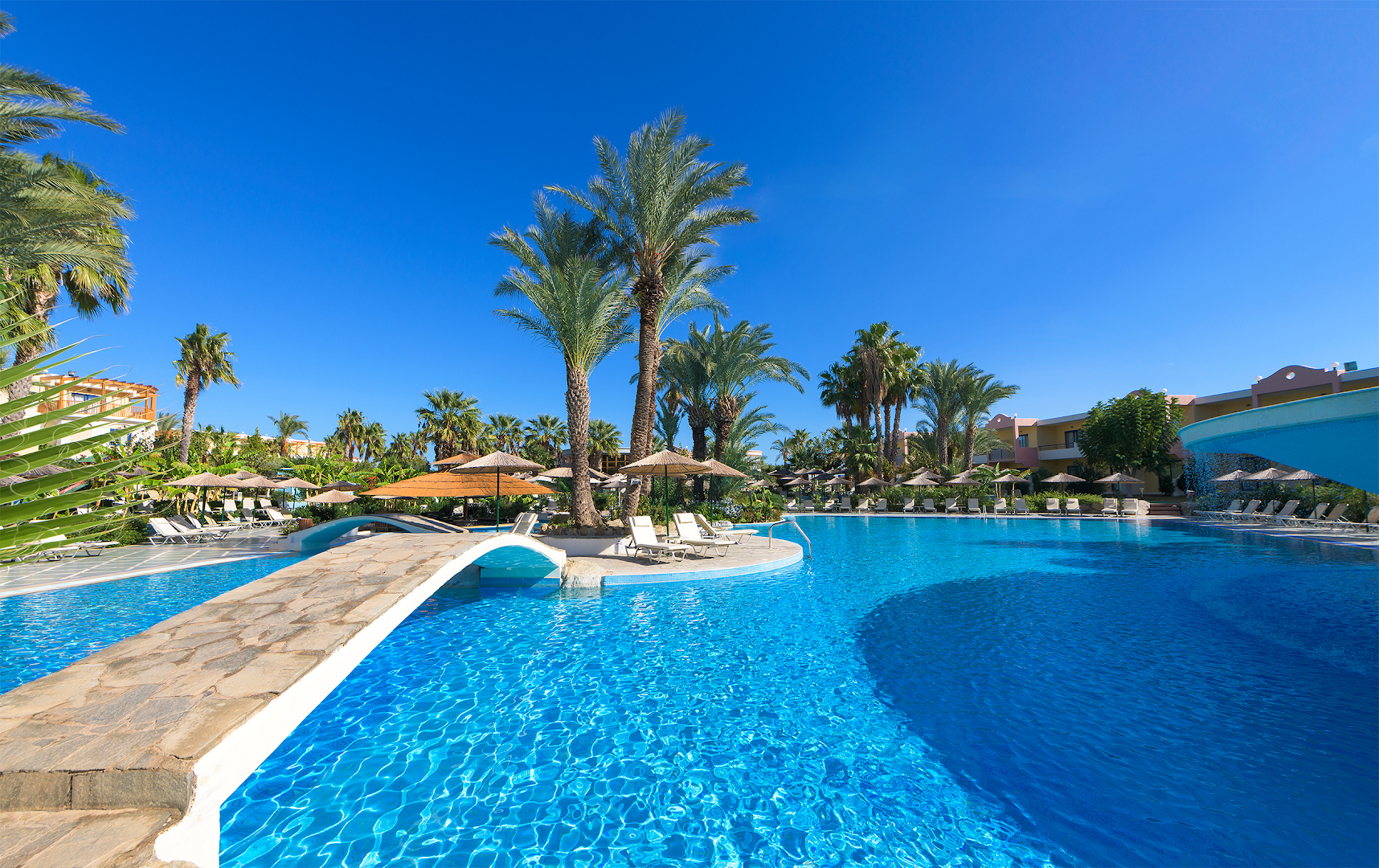 Atrium Palace Thalasso Spa Resort And Villas galvenais baseins ar akmens tiltiņu un palmām Rodas piekrastē zem skaidrām debesīm