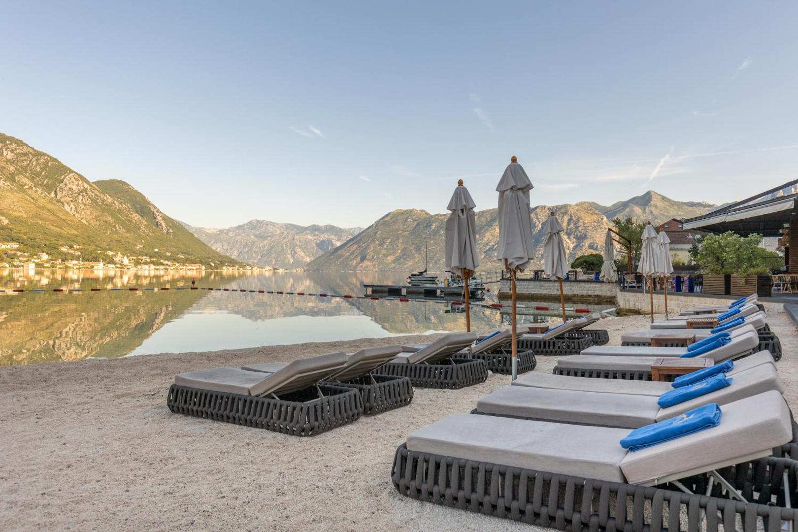 HUMA Kotor Bay Hotel & Villas privātā pludmale ar ērtiem sauļošanās krēsliem un saulessargiem pie rāmiem Kotoras līča ūdeņiem Melnkalnē