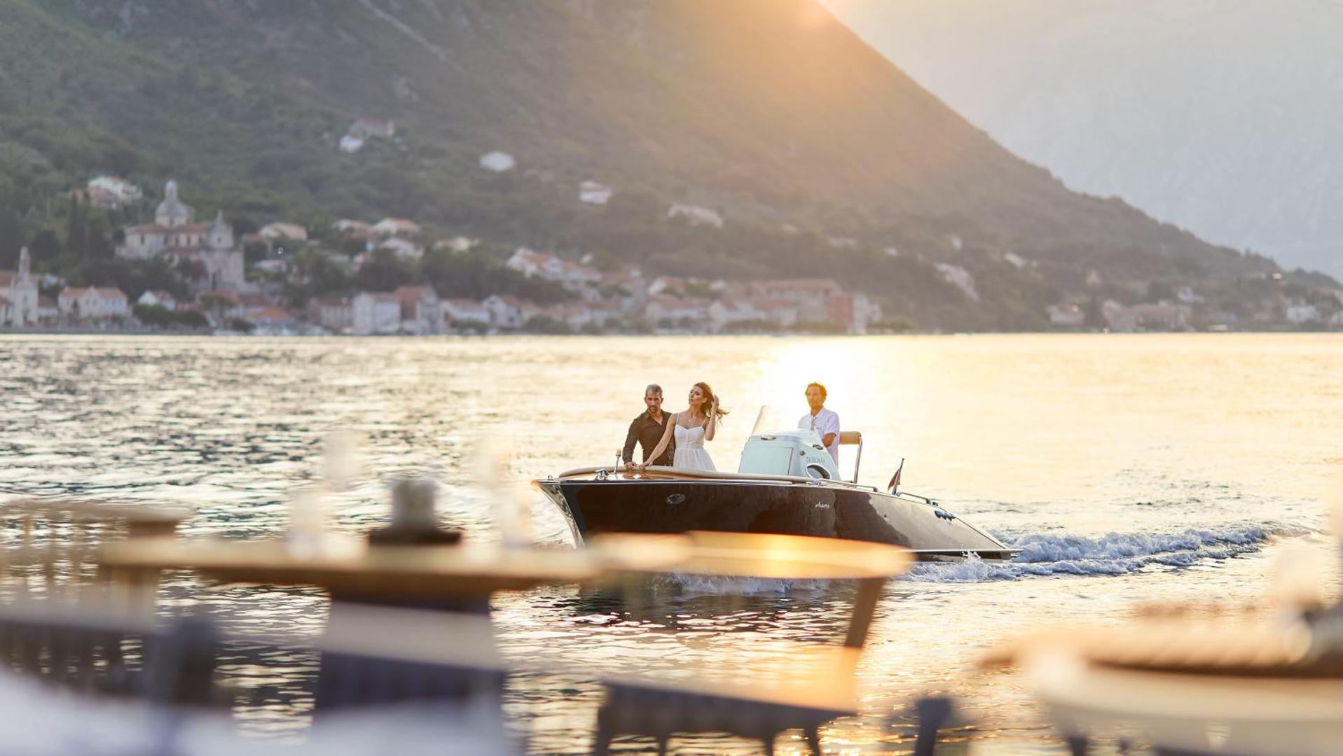 Viesi bauda braucienu ar motorlaivu saulrietā pie HUMA Kotor Bay Hotel & Villas Melnkalnē, fonā vēsturiskā piekrastes arhitektūra
