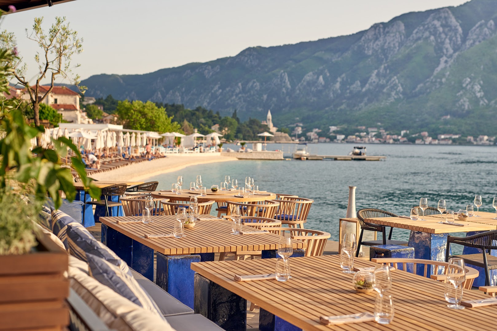 HUMA Kotor Bay Hotel & Villas restorāna terase pie jūras ar koka galdiem un skatu uz Kotoras līča kalniem