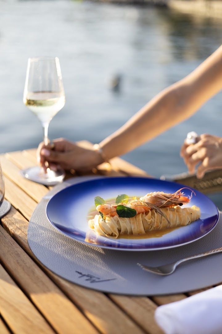 Gourmet jūras velšu ēdiens uz zila šķīvja HUMA Kotor Bay Hotel & Villas restorānā, fonā vīna glāze un līča ūdens